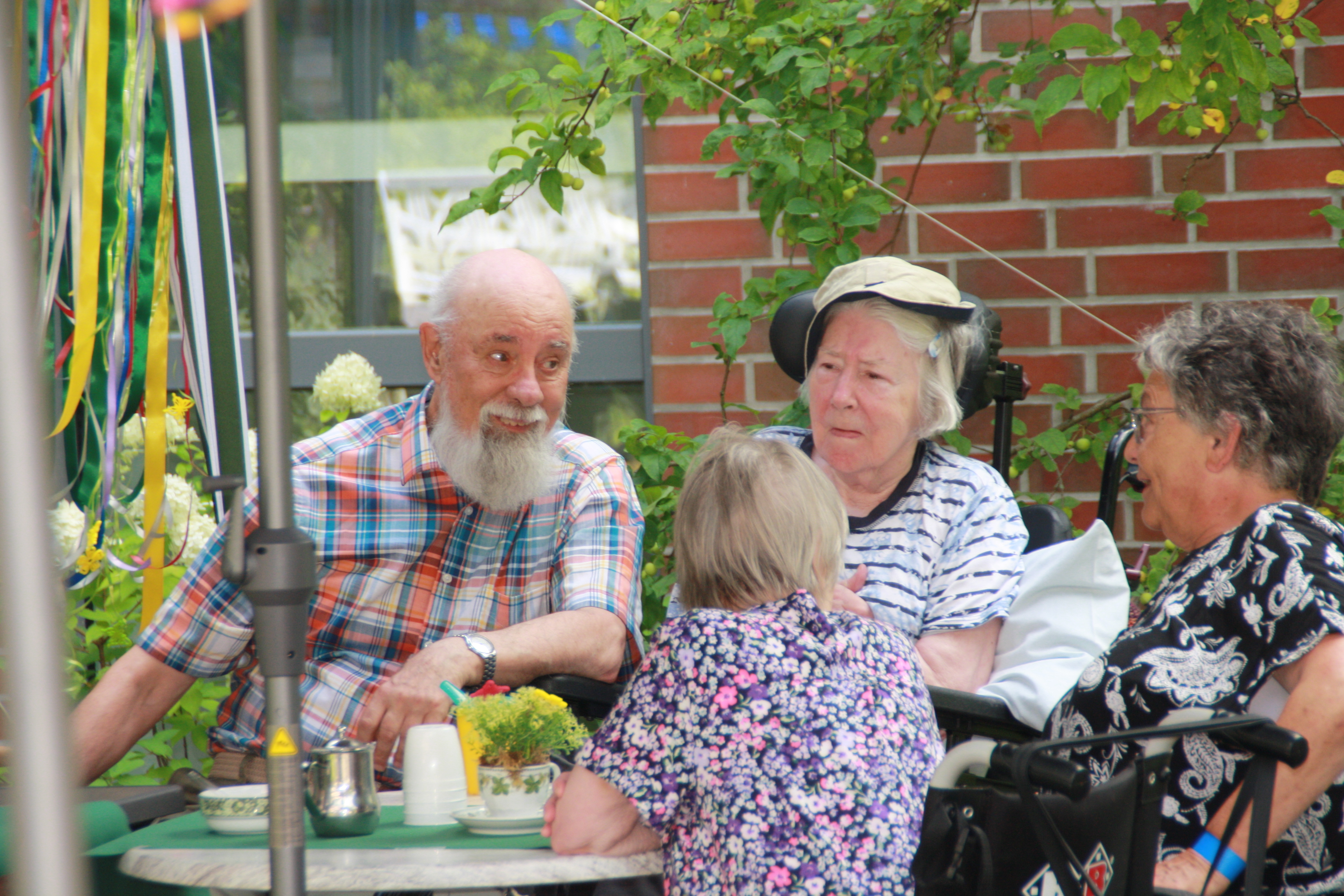 Vier ältere Menschen sitzen an einem Gartentisch und unterhalten sich. Ein Mann mit weißem Bart und kariertem Hemd sitzt drei Frauen gegenüber. Auf dem Tisch stehen Getränke und ein kleines Blumengesteck. Im Hintergrund sind eine begrünte Backsteinwand und bunte Dekorationen zu sehen.
