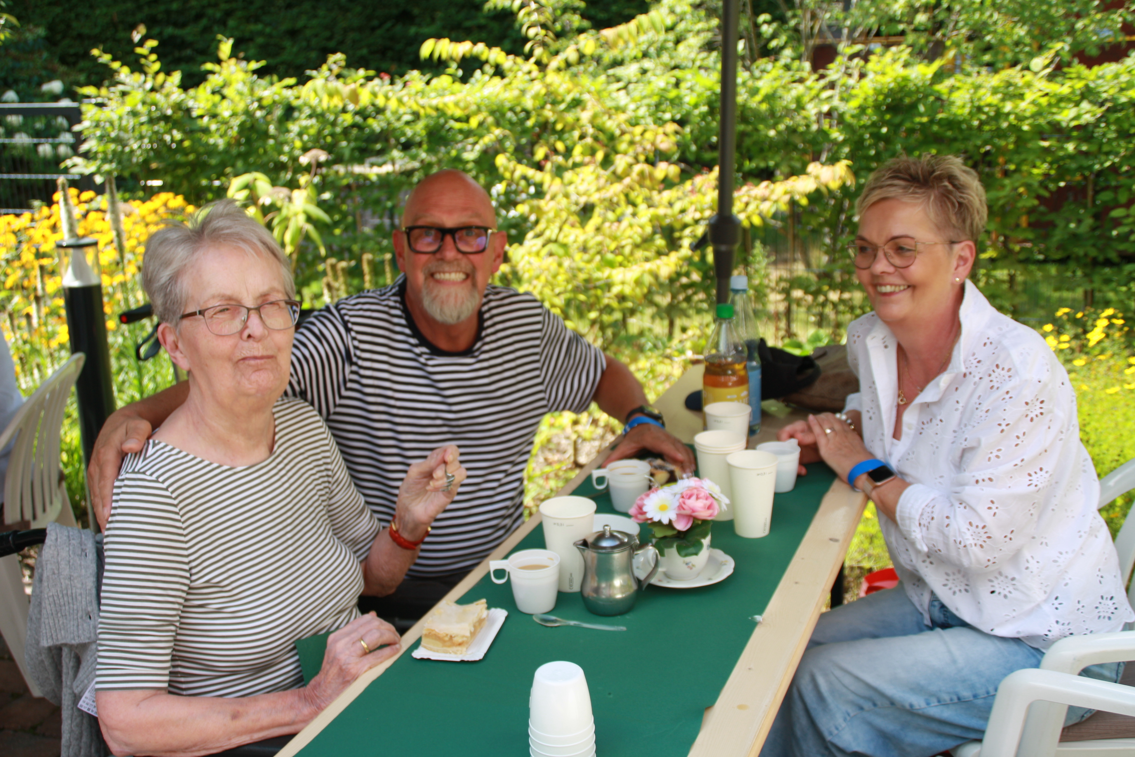 Eine ältere Frau mit Brille sitzt gemeinsam mit einem lächelnden Mann im gestreiften T-Shirt und einer Frau in weißer Bluse an einem Gartentisch. Auf dem Tisch stehen Kaffee, Kuchen und ein kleines Blumengesteck. Im Hintergrund blühen gelbe Blumen.