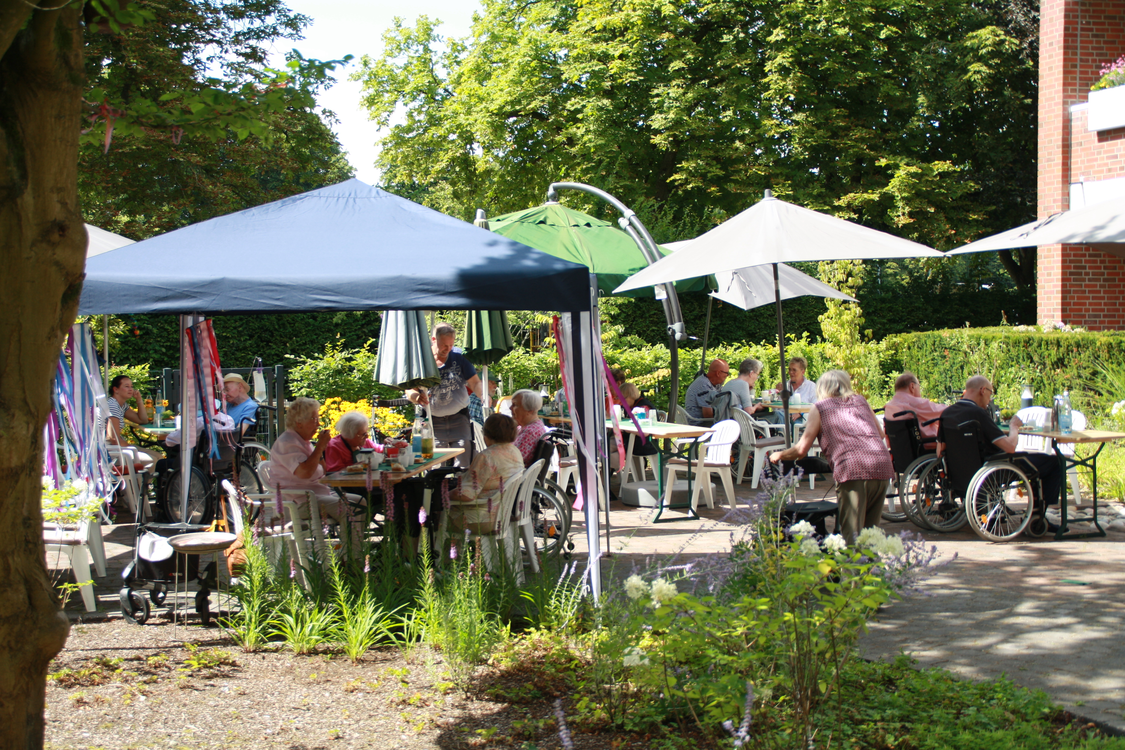 Blick auf das Sommerfest im Garten des Elisabethstifts. Zahlreiche Gäste sitzen an festlich geschmückten Tischen unter Pavillons und Sonnenschirmen. Bunte Girlanden, blühende Blumenbeete und hohe Bäume prägen die sommerliche Atmosphäre.